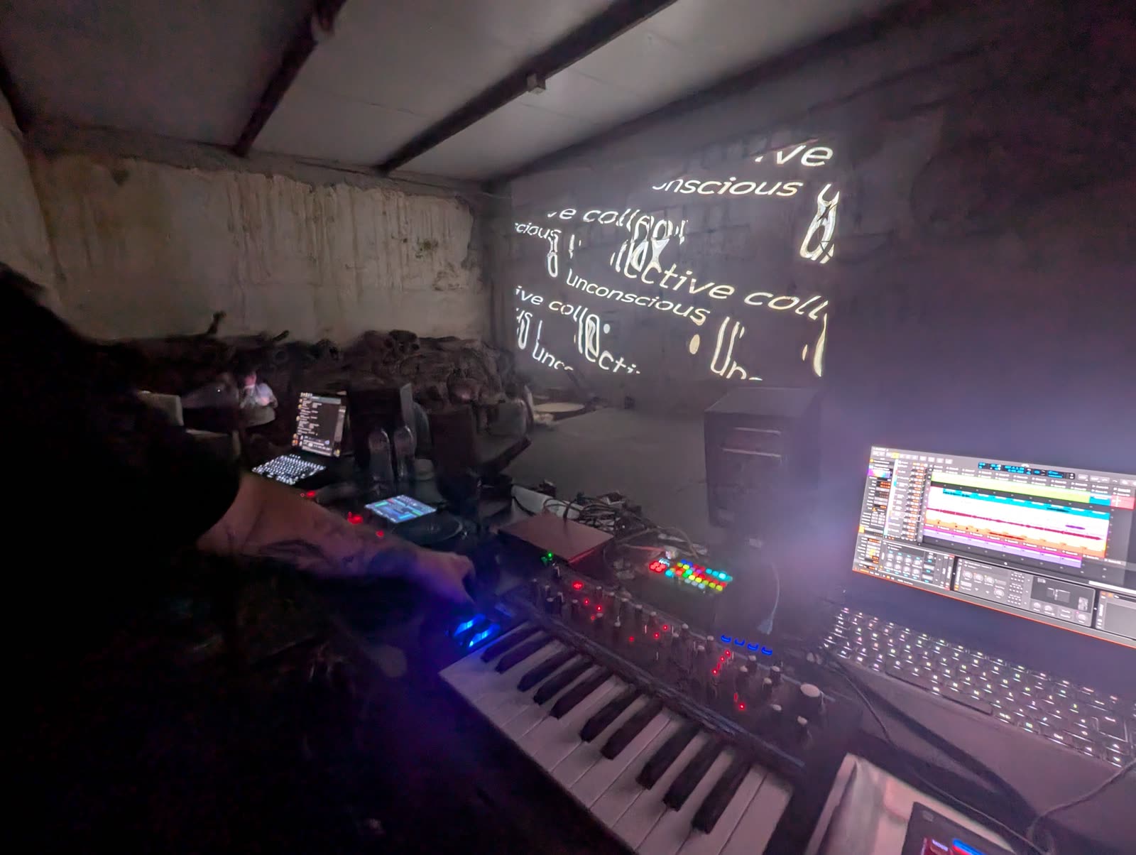Live booth with keyboard, controllers and a laptop, text projected on the wall
